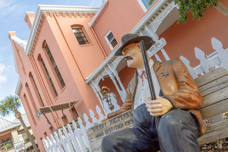 exterior shot of St. Augustine Old Jail and in the foreground is a statue of Sheriff Joe Perry sitting on a bench holding a shot gun with the words 'Sheriff Joe Perry, 1889 1897 / 1901 1919' painted on the bench